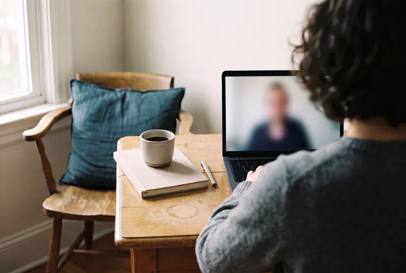 Person von hinten im Videogespräch am Laptop, daneben Notizbuch, Stift und Kaffeebecher